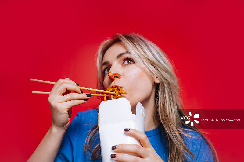 美味的面条盛宴：金发女子在红色工作室里用筷子高兴地享用亚洲炒面图片素材
