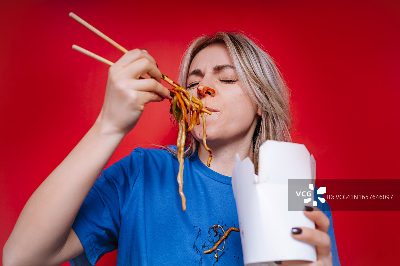 在盒子里享用酱汁面条。快乐的女人用筷子吃亚洲炒面，导致T恤沾满酱汁图片素材