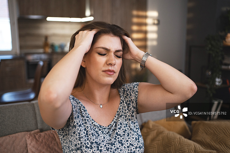 在家中经历痛苦偏头痛的年轻棕发女子，用手抱着头图片素材