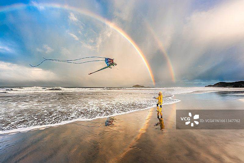 穿黄色外套的女孩在海滩上放风筝，有双彩虹图片素材