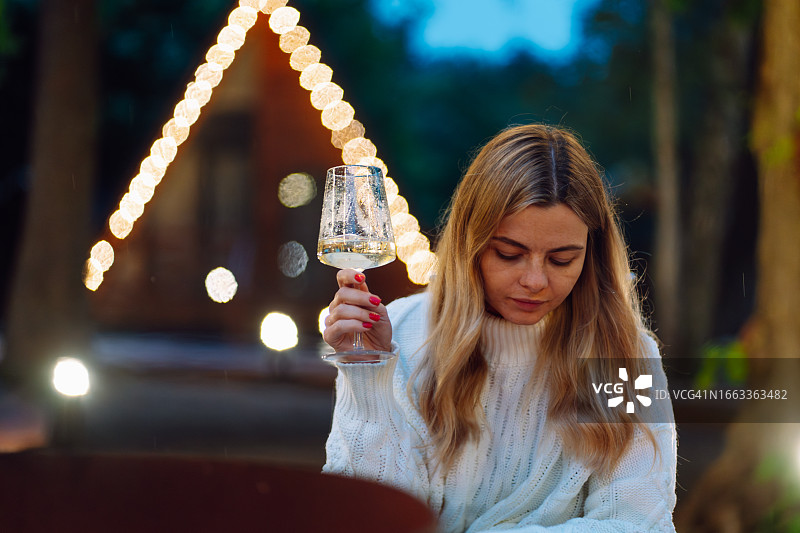 金发女人在户外放松身心，手持酒杯，背景是黄昏时分模糊的木屋图片素材