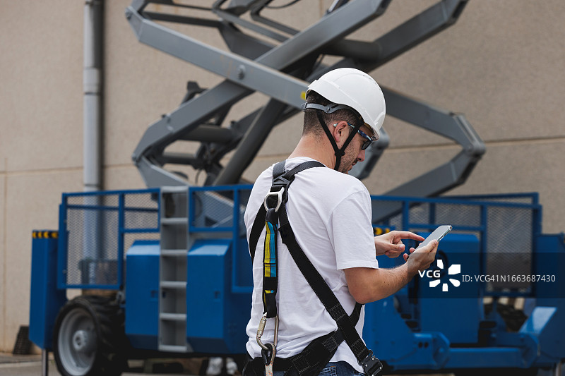 戴安全帽和安全带的工程师在升降平台附近用智能手机书写图片素材