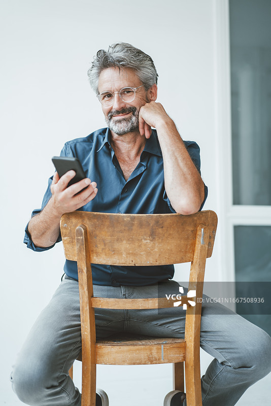 微笑的商人拿着智能手机坐在办公室的椅子上图片素材