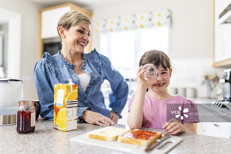 活泼女孩在厨房透过玻璃杯看妈妈。图片素材