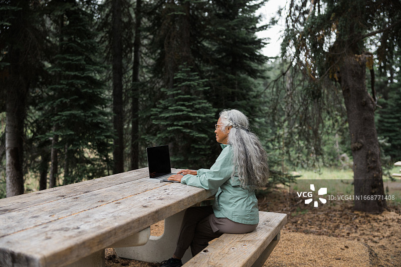 在森林野餐桌旁使用笔记本电脑的年长女性图片素材