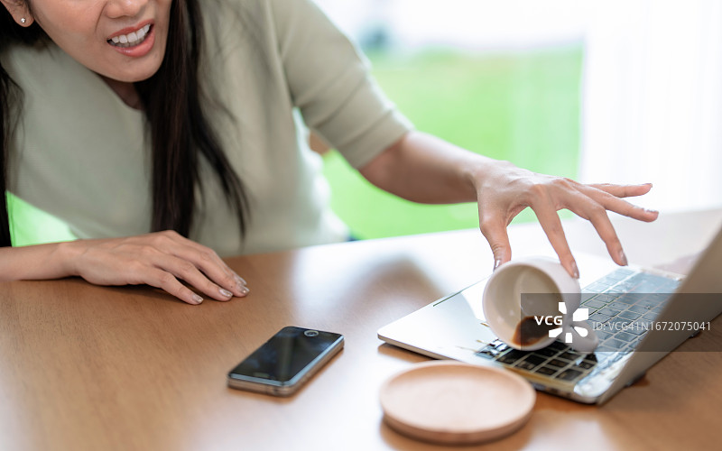 亚洲女性不小心将咖啡杯洒在笔记本电脑上，感到震惊图片素材