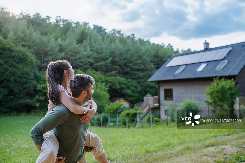 丈夫背着妻子，靠近屋顶装有太阳能电池板的家图片素材
