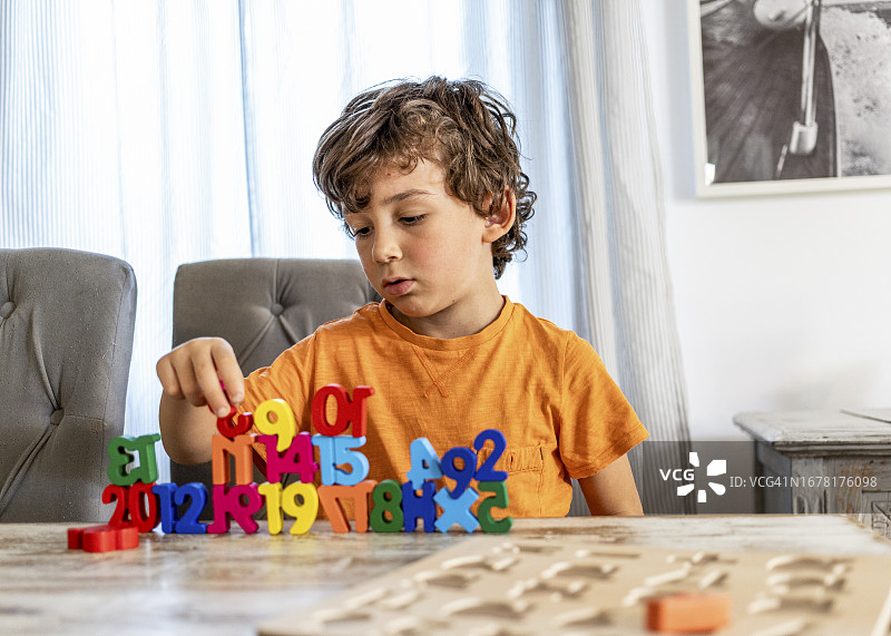 舒适的客厅氛围中,学龄前儿童和六岁卷发男孩沉浸在早晨的学习中,在餐桌上练习数字作业,周围环绕着木制数字辅助工具图片素材