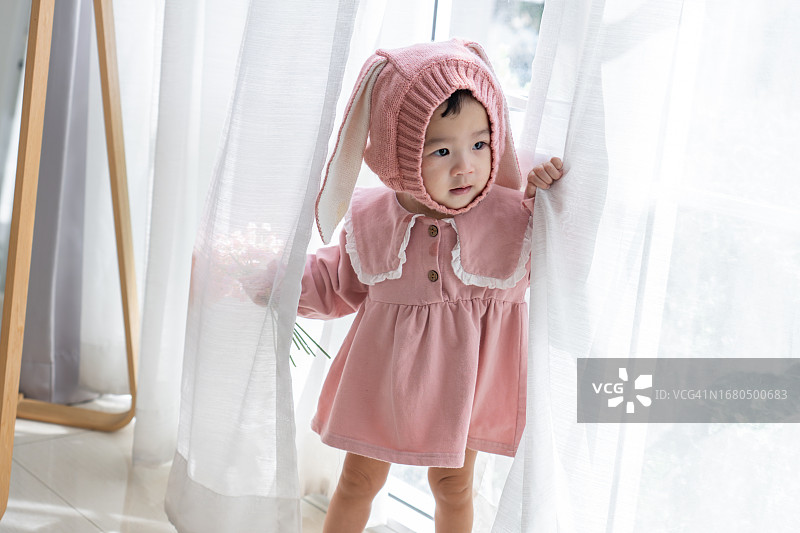 微笑的亚洲女婴站立并拿着白色透明窗帘，在窗帘后面玩捉迷藏。十个月大的可爱女孩站在白色窗帘之间。在家玩得很开心。图片素材
