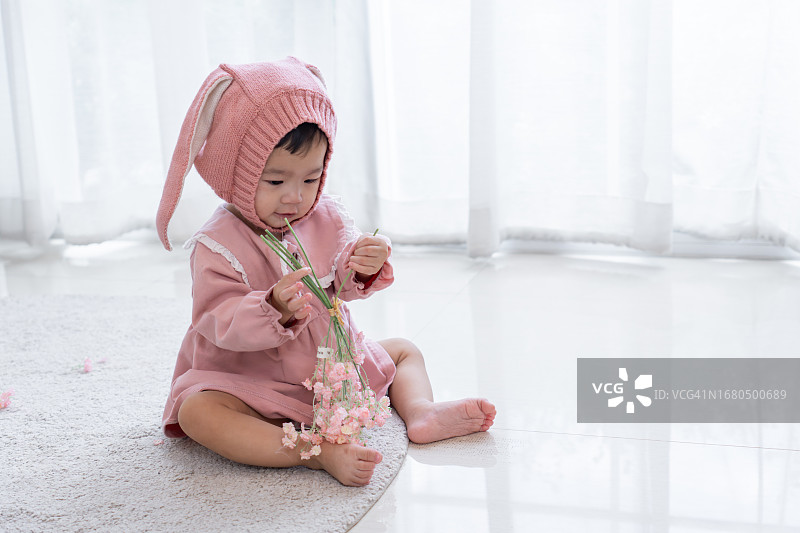 穿着粉色连衣裙和头饰的亚洲女婴在白色装饰的客厅里坐在柔软的地毯上玩着人造花图片素材