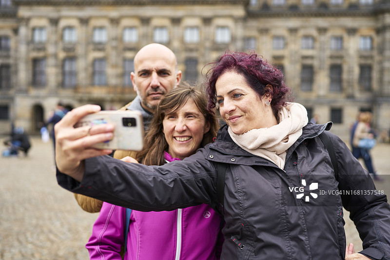 在阿姆斯特丹旅行的三人，穿着外套，拿着相机自拍，面带微笑，正面视角图片素材