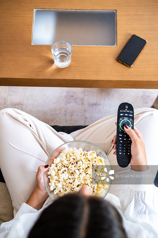 年轻女人在客厅沙发准备爆米花并用遥控器搜索电视节目图片素材