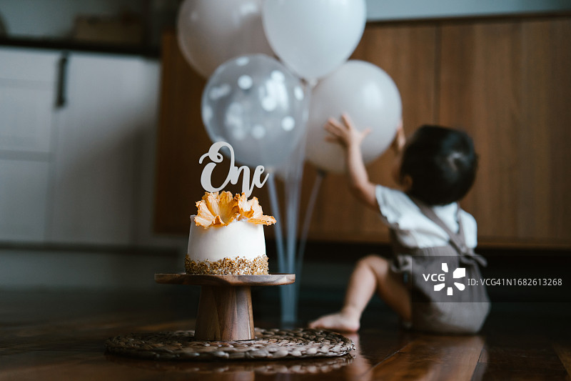 可爱的亚洲女宝宝庆祝一周岁生日，在客厅地板上玩气球，焦点在木制蛋糕架上的生日蛋糕，生日庆祝主题图片素材