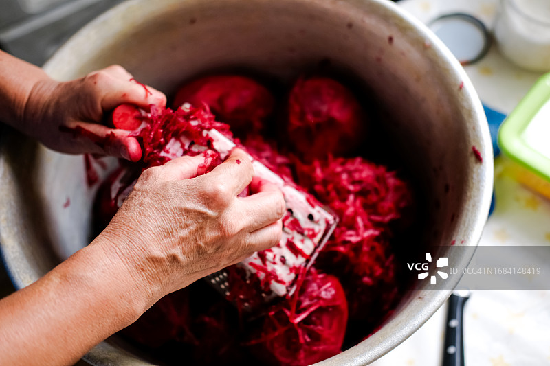 一位57岁妇女用磨碎机在一个大容器上削甜菜的手部特写，俯视图。准备甜菜库存图片素材