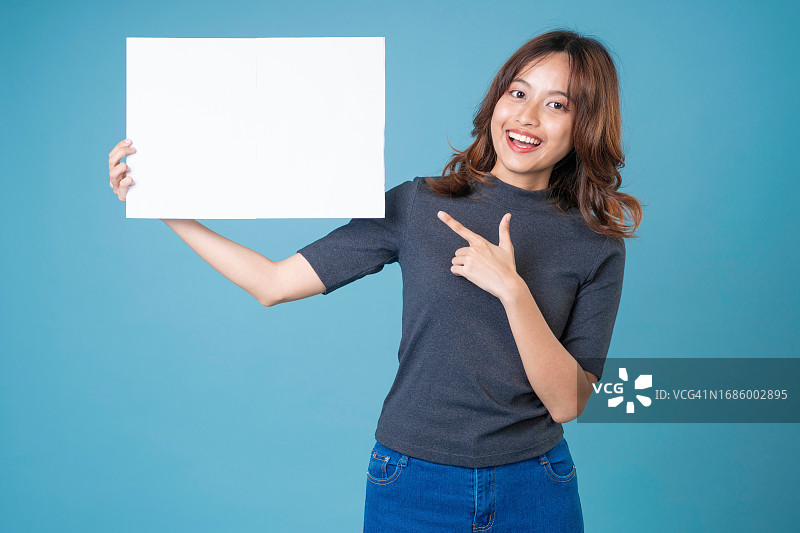年轻女人拿着浅蓝色背景上的空白板，亚洲女人展示并拿着空白白色广告牌图片素材