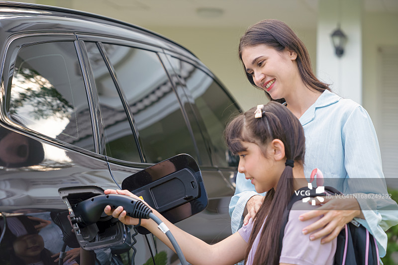 年轻的母亲和女儿在院子里给电动汽车充电，准备去上学图片素材