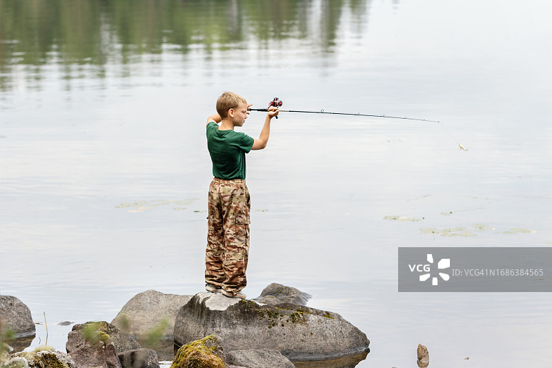 儿童渔夫 - 小男孩拿着钓鱼竿站在石头上，在飞蝇钓鱼期间将钓鱼饵扔入水中图片素材