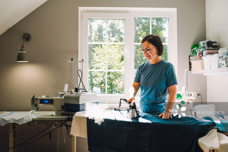 缝纫室内熨烫：戴眼镜的棕发裁缝在明亮的工作室内熨烫面料图片素材