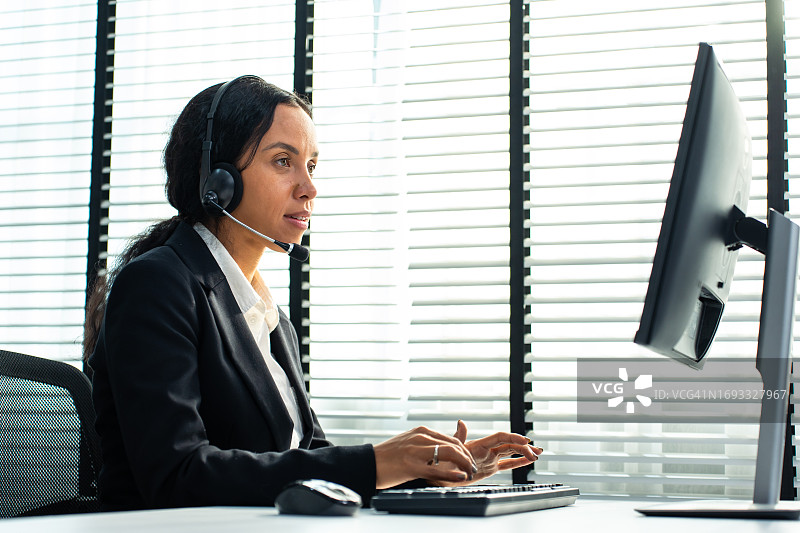 在公司办公室工作的拉丁裔美女客服中心员工。有魅力的年轻员工坐在桌旁，戴着耳机，使用笔记本电脑与客户沟通图片素材