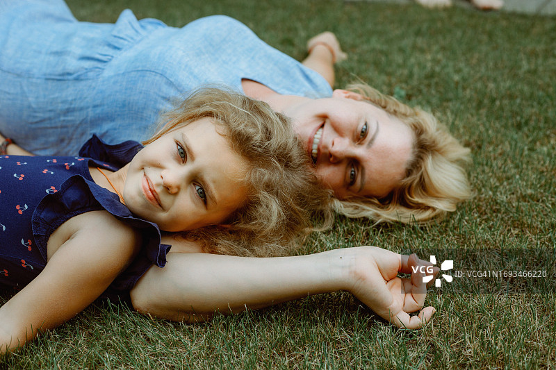 快乐的中年女士穿着蓝色连衣裙躺在草坪上，旁边躺着穿着深蓝色连衣裙的漂亮小女孩图片素材