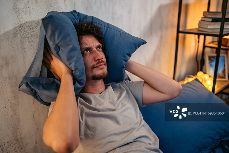 晚上在卧室里用枕头捂住耳朵的愤怒年轻人图片素材