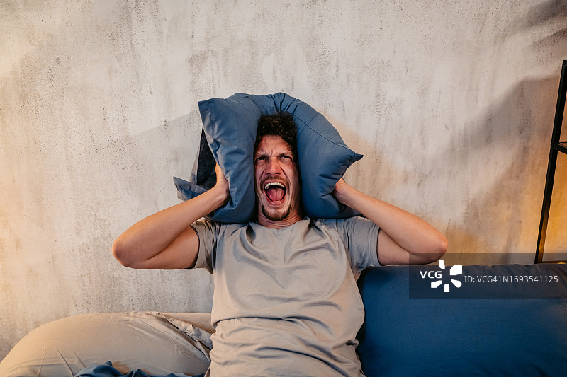 晚上在卧室里用枕头捂住耳朵的愤怒年轻人图片素材