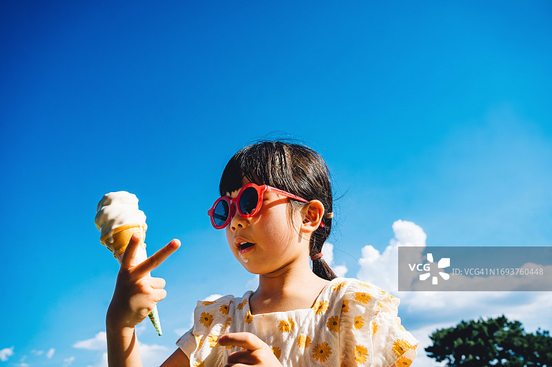 阳光明媚的夏日，可爱的亚洲小女孩戴着红色太阳镜在户外吃融化的冰淇淋图片素材