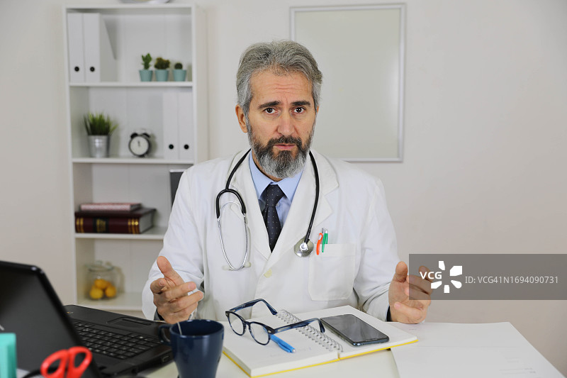 戴眼镜的白发男医生在办公室办公桌前使用无线笔记本电脑打字，看着镜头。科技与医学概念图片素材