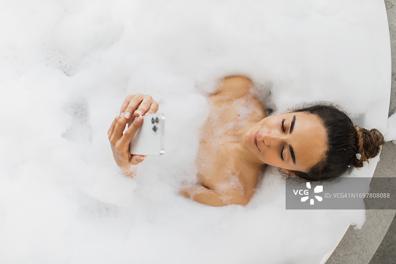 年轻漂亮的女人躺在满是泡沫的浴缸里，用手机看电影图片素材