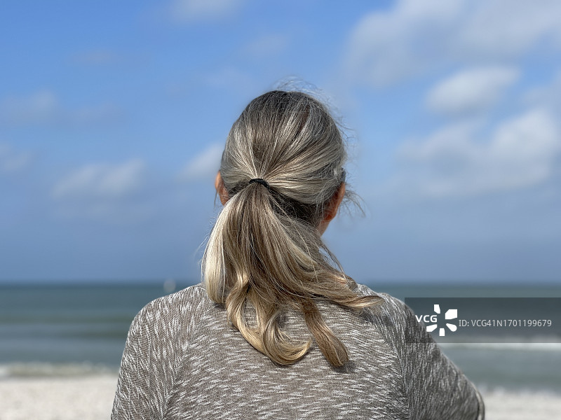 在海滩上眺望大海的扎马尾年长女性，背景是海水、蓝天和白云图片素材