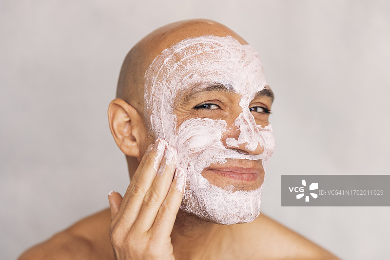 在白色背景下往脸上涂抹面霜的微笑光头成熟男人肖像图片素材