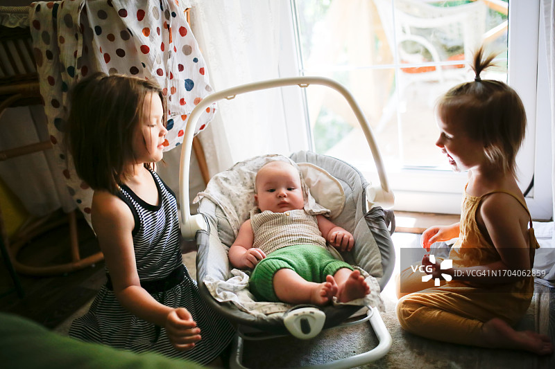 姐妹俩在家看着在电动摇椅上的胖乎乎的弟弟图片素材