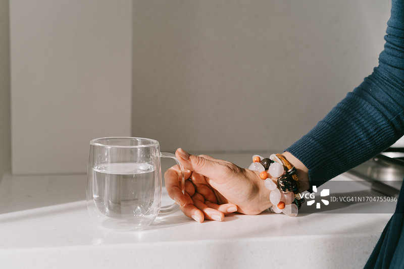 女人站在厨房柜台旁，拿着一个透明的马克杯或一杯水。身体水分平衡、脱水问题、健康饮食、服用营养补充剂和维生素、解渴的概念图片素材