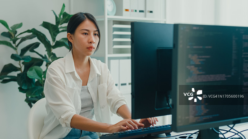 年轻的亚洲女性软件开发人员使用电脑编写代码，坐在办公桌前，多个屏幕在办公室工作。程序员开发。图片素材