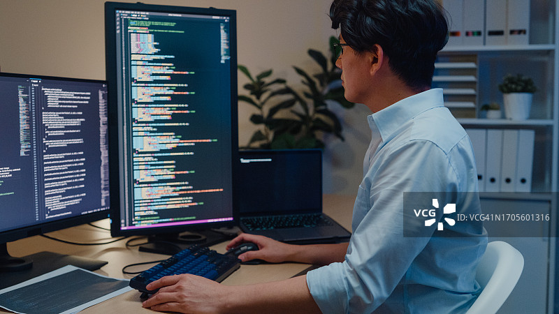 年轻的亚洲男性软件开发人员使用电脑编写代码，坐在办公桌前，在办公室里使用多个屏幕工作。程序员开发。图片素材