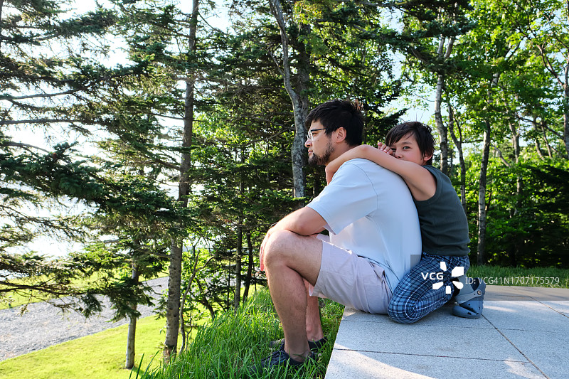 男人坐着望着远方，他的儿子从背后拥抱他。图片素材