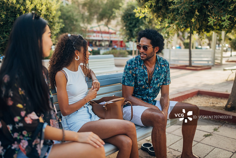 不同种族的朋友们在阳光明媚的日子里坐在公园的长椅上聊天，分享度假见闻图片素材