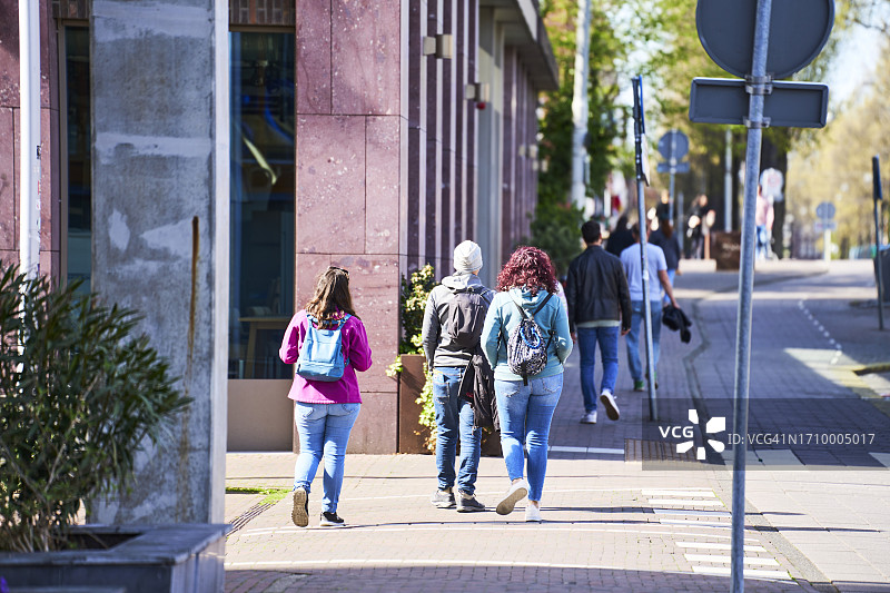 一群人背着背包和外套在建筑物外的人行道上行走的后视图图片素材