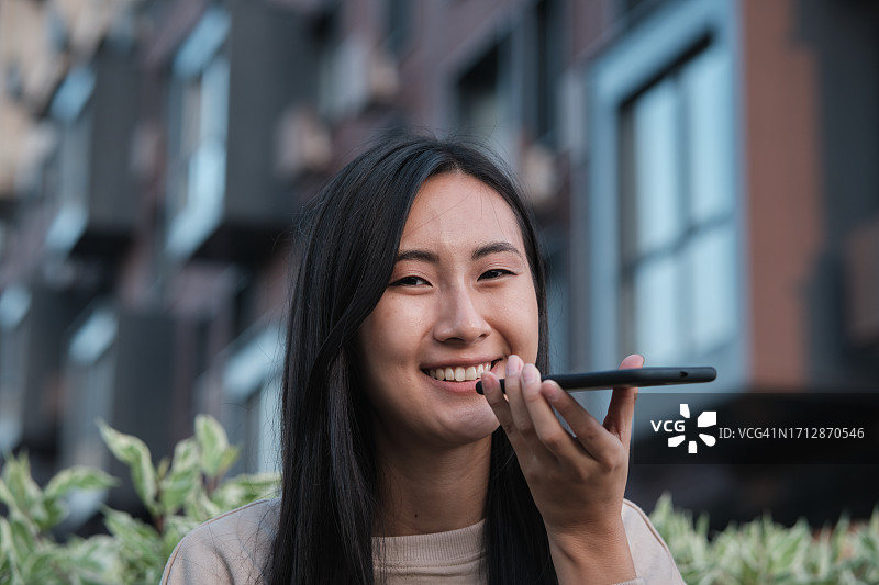 快乐的亚洲女人在户外使用免提电话发送语音邮件，千禧一代韩国女孩拿着智能手机发送语音信息图片素材