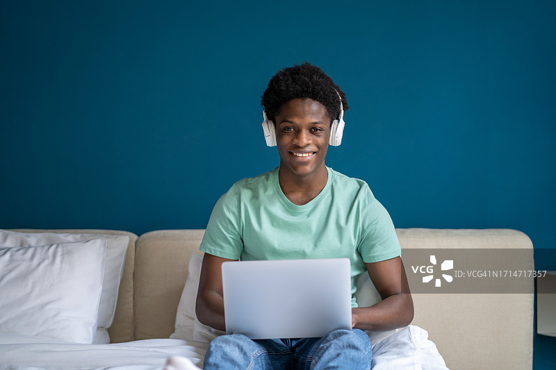 微笑的非洲裔美国男子戴着无线耳机坐在床上，膝盖上放着笔记本电脑，看着镜头图片素材