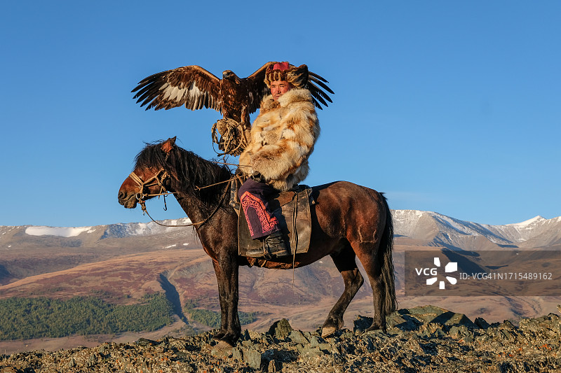 一位哈萨克族驯鹰人坐在阿尔泰山的马背上，手臂上架着一只鹰图片素材