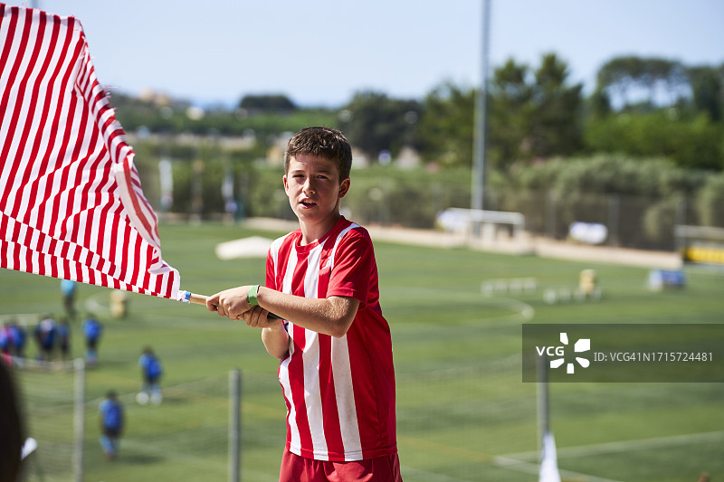 身穿运动服站在体育场看台上，手持旗帜，看向镜头的男孩肖像（正面）图片素材