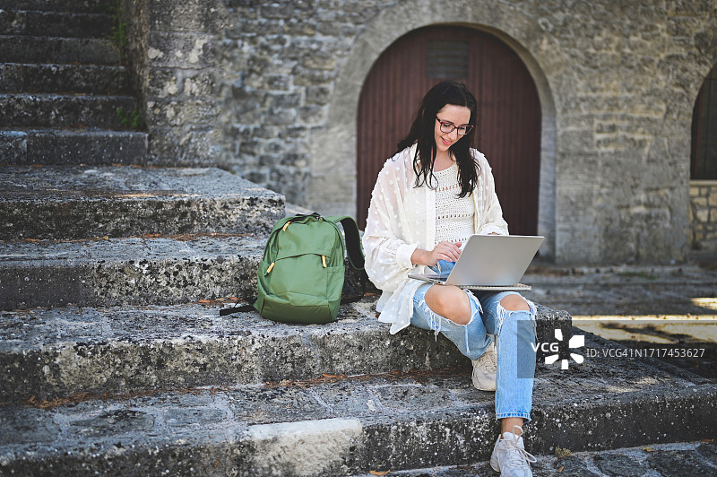 背着背包在楼梯上用笔记本电脑工作的漂亮年轻女人图片素材