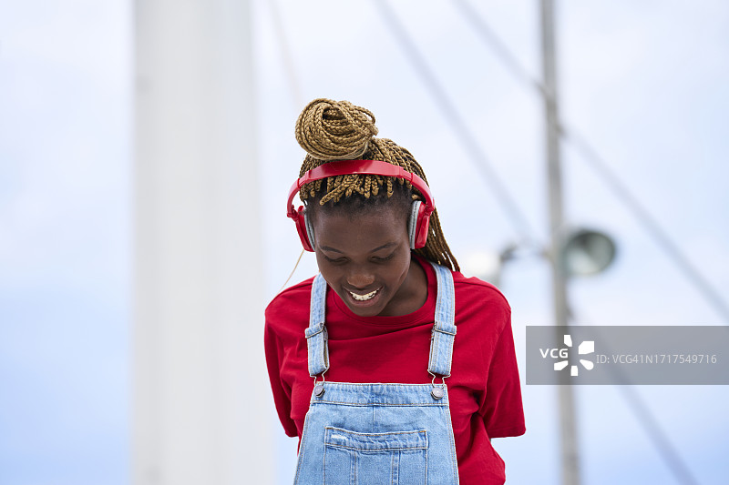 戴着耳机、低着头微笑的深色皮肤女孩，头顶盘着发髻，金发上编着辫子，背景虚化图片素材