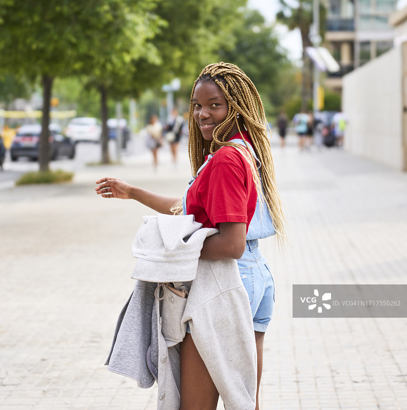 年轻女子站立着，侧身对着镜头，左臂上搭着一件外套，背景是树木和行人图片素材