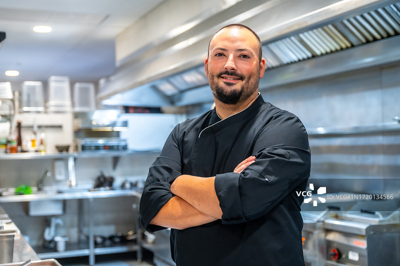 餐厅厨房内身穿黑色制服的厨师画像，厨师面带微笑，双臂交叉图片素材
