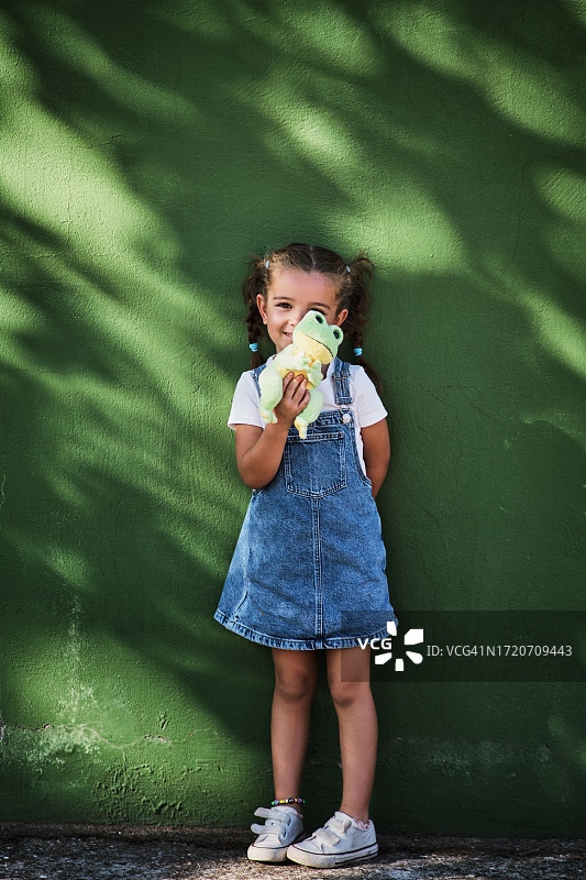 抱着毛绒玩具青蛙，靠在绿色墙壁上的小女孩肖像图片素材