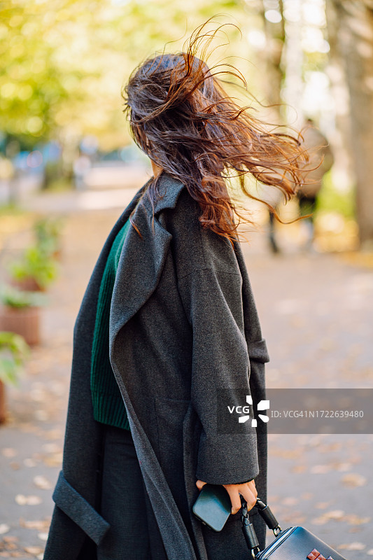 在秋天的公园里甩头发和转身的棕发女子。穿着现代外套、拿着智能手机在公园里漫步的女子，看不清面容，看向远离相机的方向图片素材
