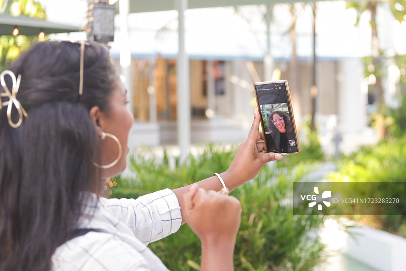 在热带气候城市的户外零售场所，一位千禧一代拉丁裔女性在手机上与朋友进行视频通话图片素材