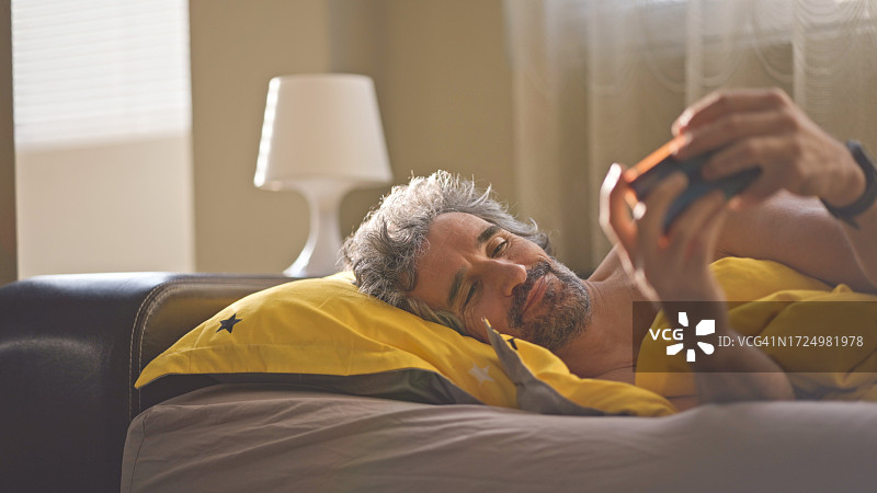 中年男人醒来后在床上用手机浏览社交媒体。生活剪影。图片素材